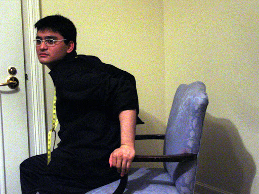 Josh tests the posing chair in the corner of the room. Measuring tape is wrapped around his neck. Josh tests the posing chair in the corner of the room. Measuring tape is wrapped around his neck.